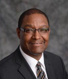 Dennis Mitchell, wearing a dark gray suit, white dress shirt, and glasses, smiles into the camera in front of a charcoal gray background