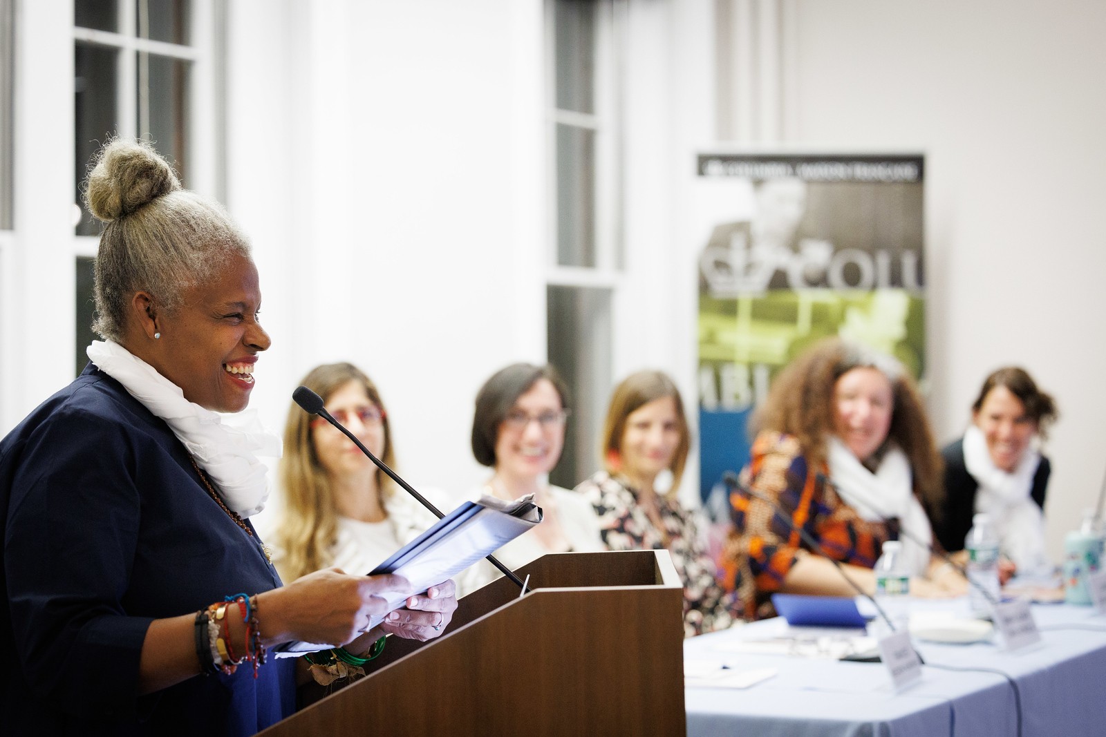 Josefina Báez and panelists Josefina Baez standing at podium holding book, with five women seated out of focus