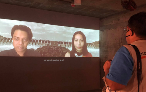 Naeem Mohaiemen + Samari Chakma, Bor Porong: Autobiography of the Drowned project on indigenous people of southeast Bangladesh. Installation view at Chobi Mela 0, Drikpath Bhavan, 2021. Photo: Suborna Morsheada Photo of video installation with faces of Naeem Mohaiemen and Samari Chakma on the screen