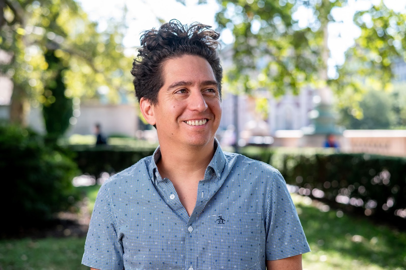 Photo of Daniel Alarcon wearing blue short sleeved shirt smiling and looking to the side with trees in the background