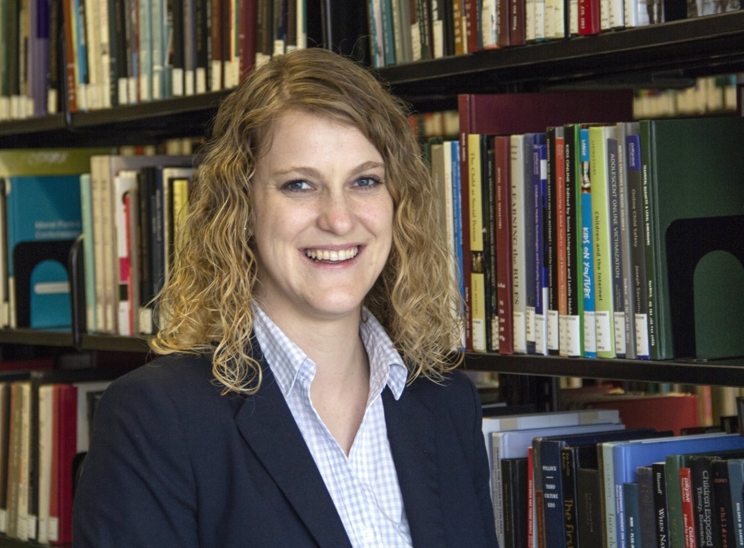 Photo of Alissa Davis with curly blonde hair wearing a white shirt and dark jacket standing in front of bookshelf