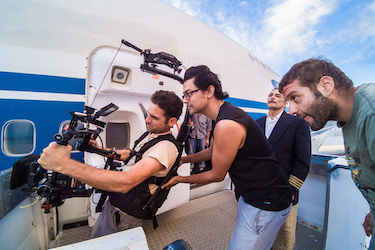 Filming Tripoli Cancelled at Ellinikon Airport, Athens, Greece, 2016. The film was a finalist for Britain's Turner Prize (2018). Ellinikon Airport has since been demolished, as part of a luxury real estate redevelopment project. Photo: Dimitris Parthimos Naeem filming Tripoli Cancelled, standing behind a camera man with two other men looking on. They are standing next to a blue and white airplane.