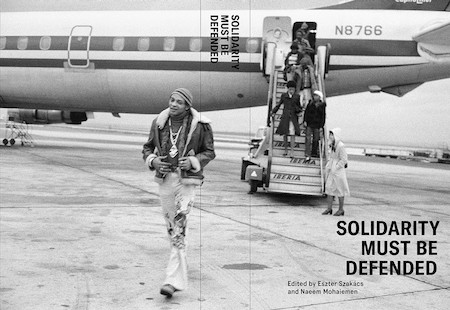 Black and white Book cover of Solidarity Must Be Defended featuring a photo of a man walking off a plane while others are behind him going down the steps.
