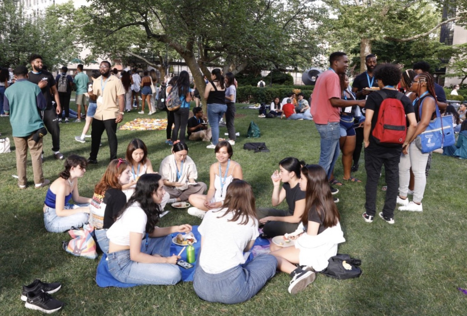 Photo of several students seated in a circle on the grass; in the background, small groups of students stand talking
