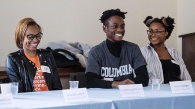 Photo of three panelists smiling at table. One wearing black jacket and orange shirt, one wearing a Columbia Tshirt and long sleeved shirt, and one wearing a white shirt opened over a black shirt.