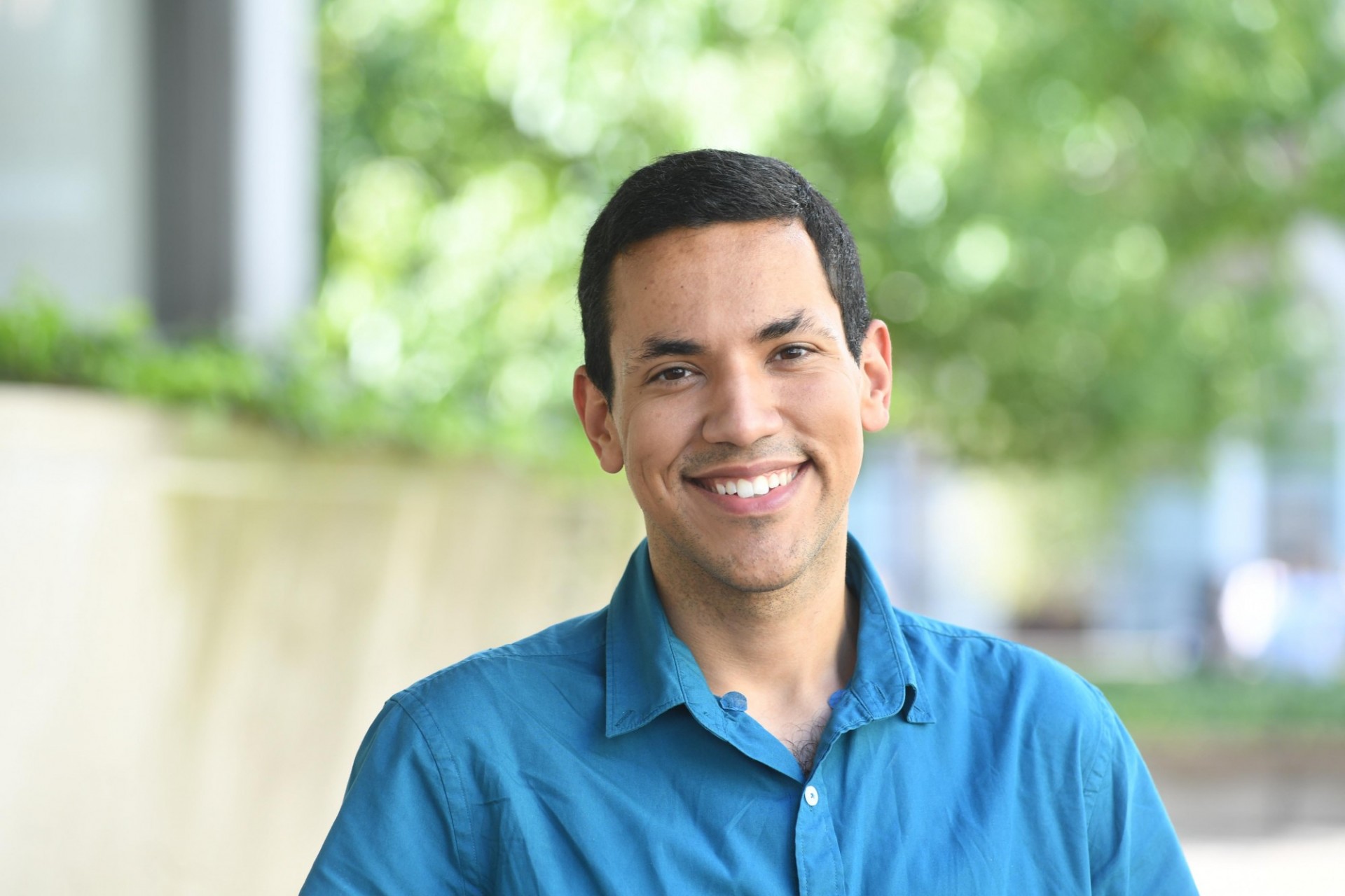 Portrait of Brandon Agosto, a young latino male with short hair, wearing a blue shirt against a green outdoor faded background