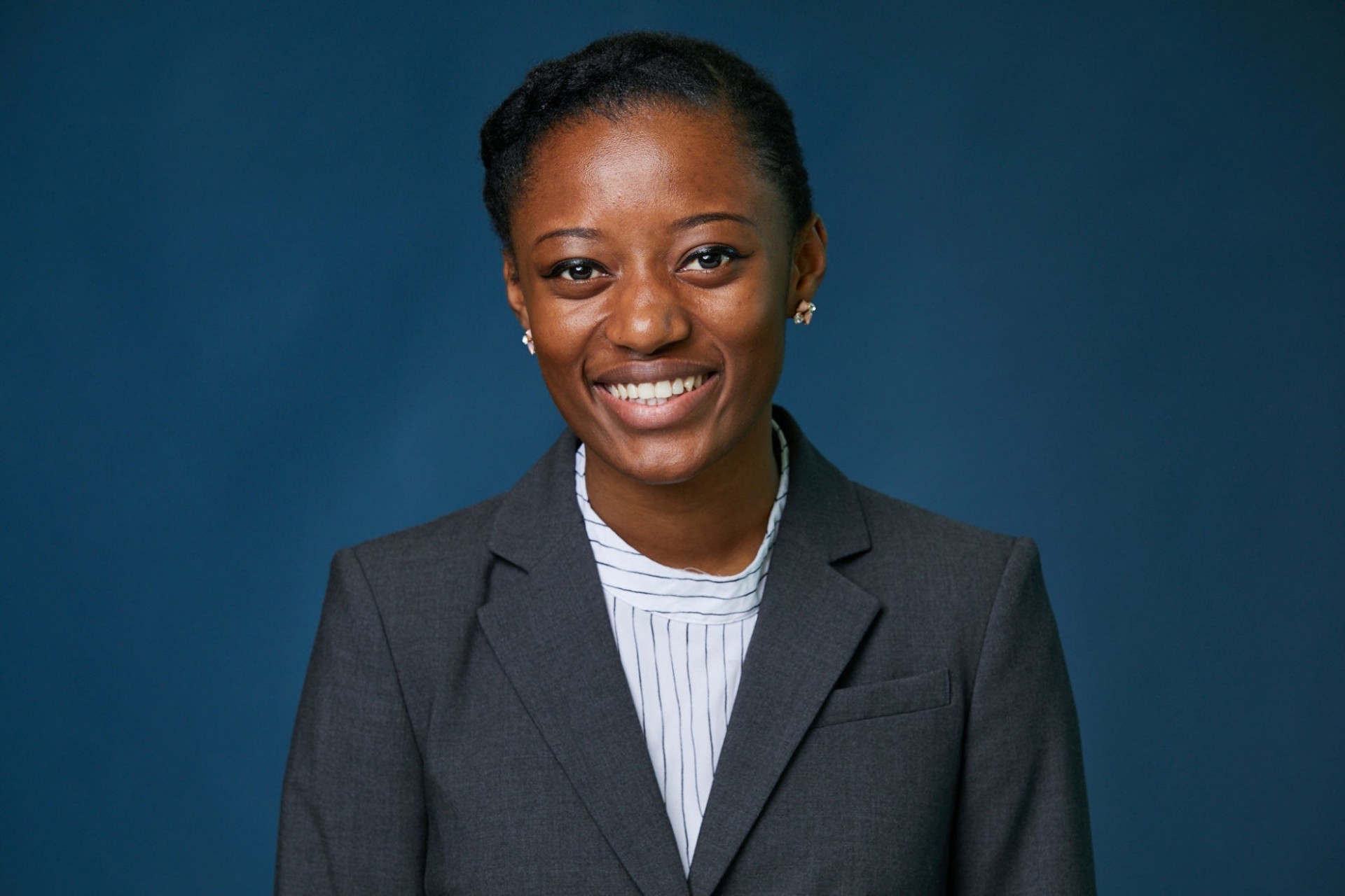 Portrait of Daniela Bushiri, a young female with black hair pulled back, dressed in a grey suit and white stripped shirt, against a dark blue background