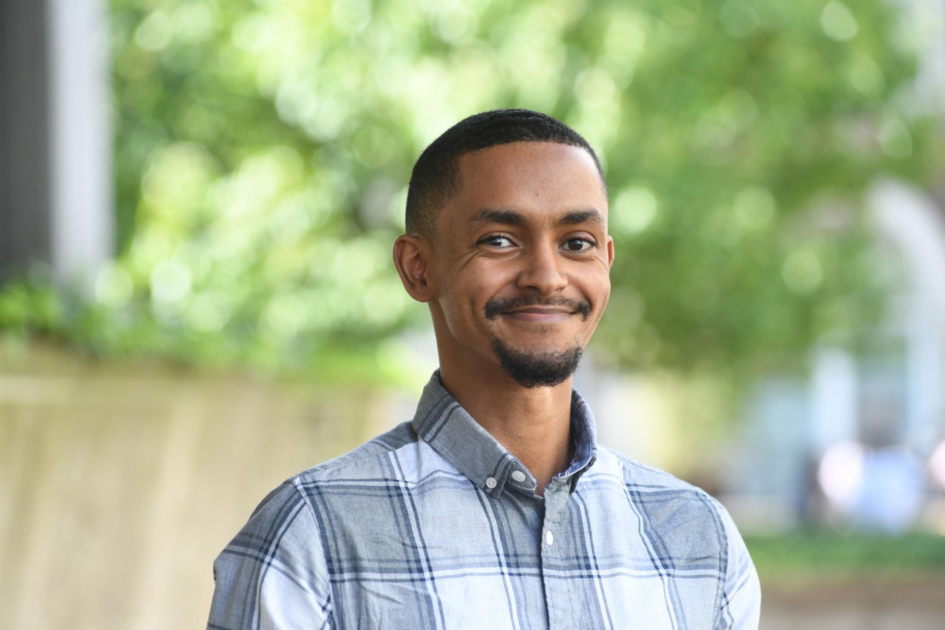 Portrait of Betselot Wondimu, a young male with short hair, a short goatee beard and mustache, wearing a white and blue pleated shirt against a green outdoor faded background