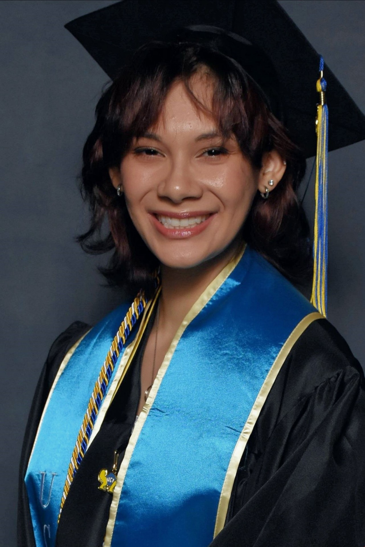 Portrait of Emily Barragan, a young woman with brown hair wearing black cap and gown with blue and gold regalia on a grey background