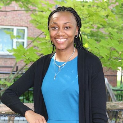 Portrait of Kevonyah Edwards, a young woman with long braided hair, dressed in a blue shirt and a black sweater, against a green background and a building