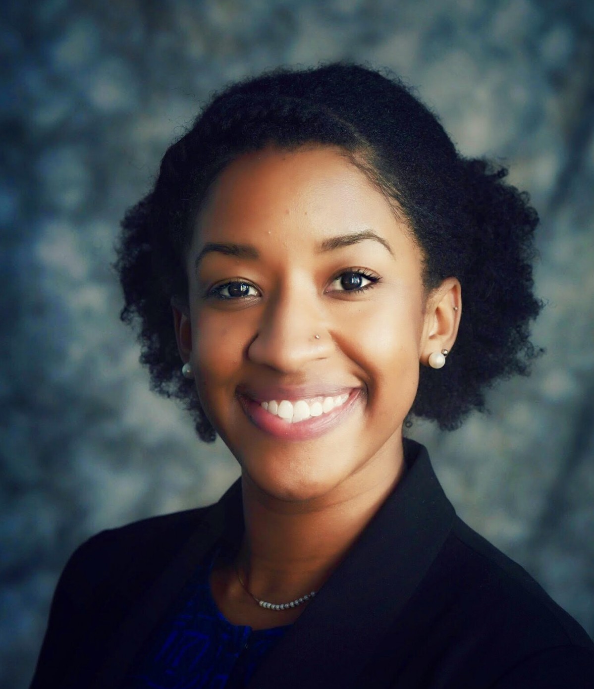 Headshot of Kristen Barnes with dark curly hair wearing a black suit in front of a blue background