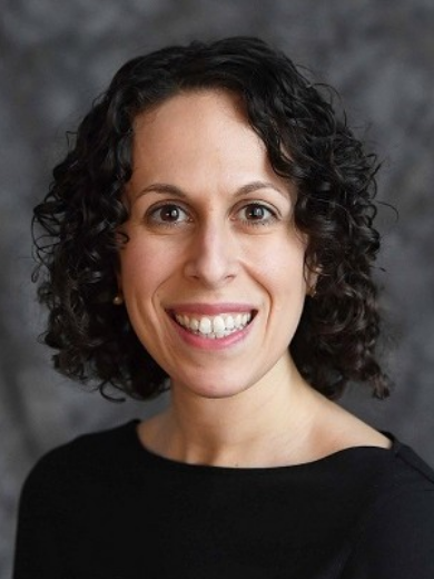 Headshot of smiling Rebecca Balkin wearing a black shirt, with curly dark hair in front of a gray background.