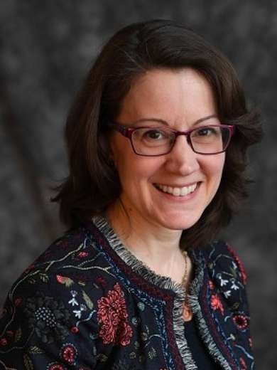 Head shot of Kristin Carnahan wearing a multicolored jacket and dark red glasses, smiling in front of gray background