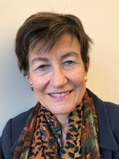 Photo of Paula Goodman with short brown hair and wearing a black jacket and multicolored scarf, smiling in front of off-white wall