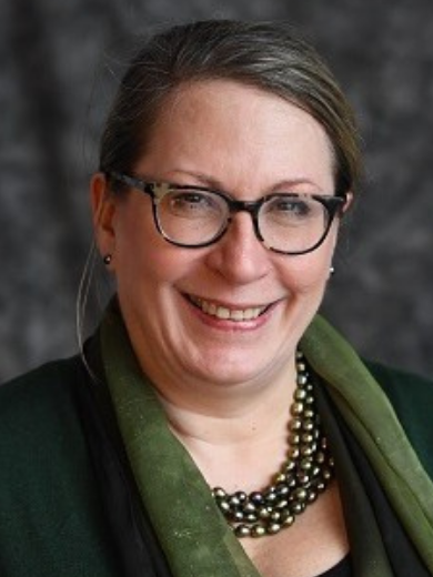 Head shot of Alice Lesman wearing glasses and a green necklace and scarf with a black jacket