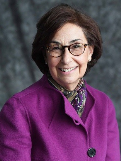 Marcia Roesch wearing a magenta jacket with short straight hair, smiling in front of a gray background
