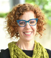 Photo of Jen Leach, with red curly short hair and turquoise glasses, wearing a black shirt and a green scarf