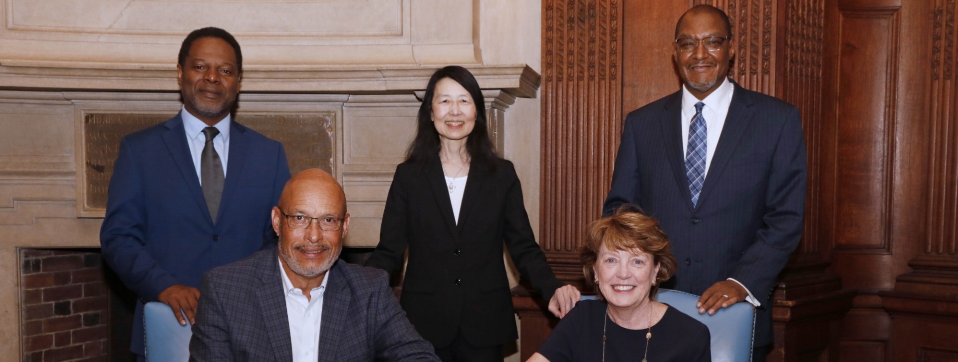 A man and woman sit at a table with documents in front of them. Three people in suits stand behind them.