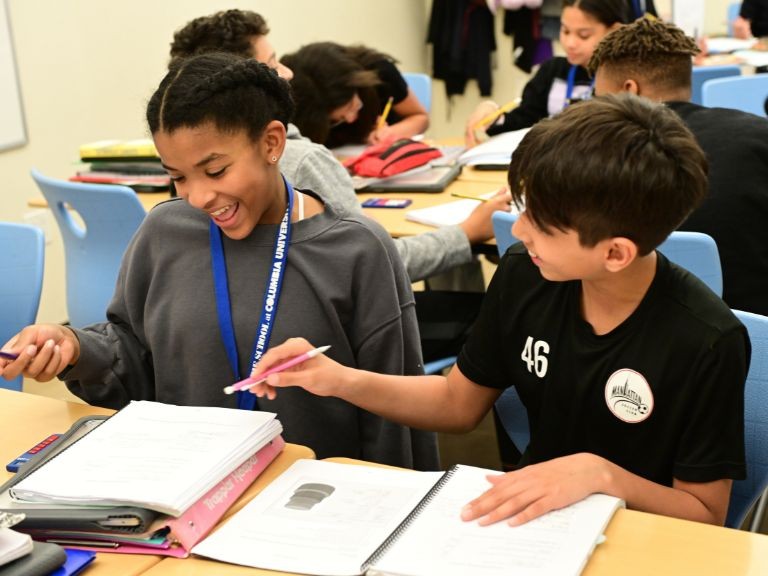 Diverse middle school classroom. Students are filling out worksheets. Young boy seated next to girl points at her notebook and they both smile.