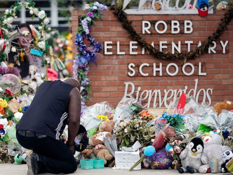 Man on one knee grieving in front of Robb Elementary School littered with flowers and wreaths after school shooting. 
