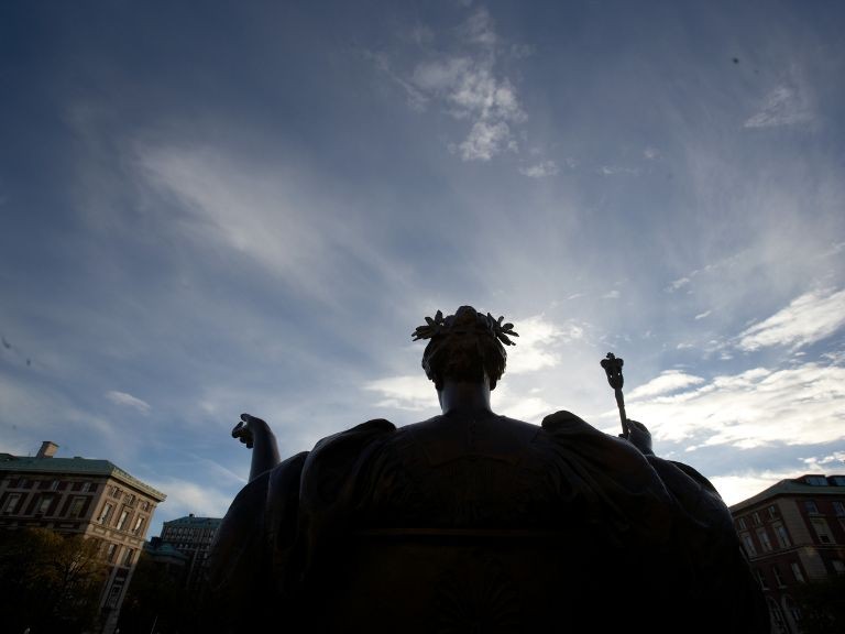 Statue of seated woman, wearing a robe and wreath, with arms outstretched, against a partly cloudy sky.