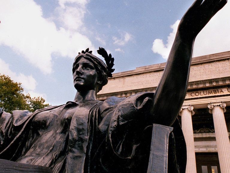 Bronze statue of woman with outstretched arms, seen from below in front of a white building with columns and a blue sky