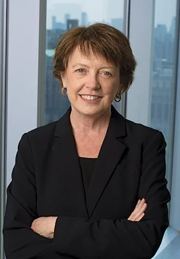 Mary C. Boyce wears black blazer and has arms crossed, standing in front of a window with a view of New York City