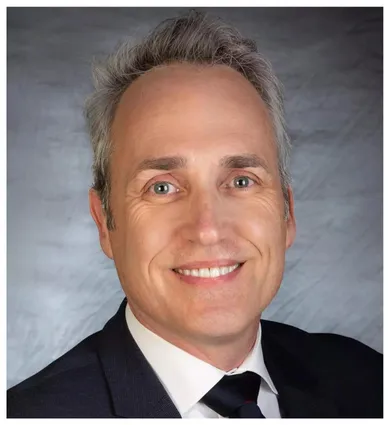 Head shot of Walter Bockting, wearing a black jacket and tie, in froont of a gray background
