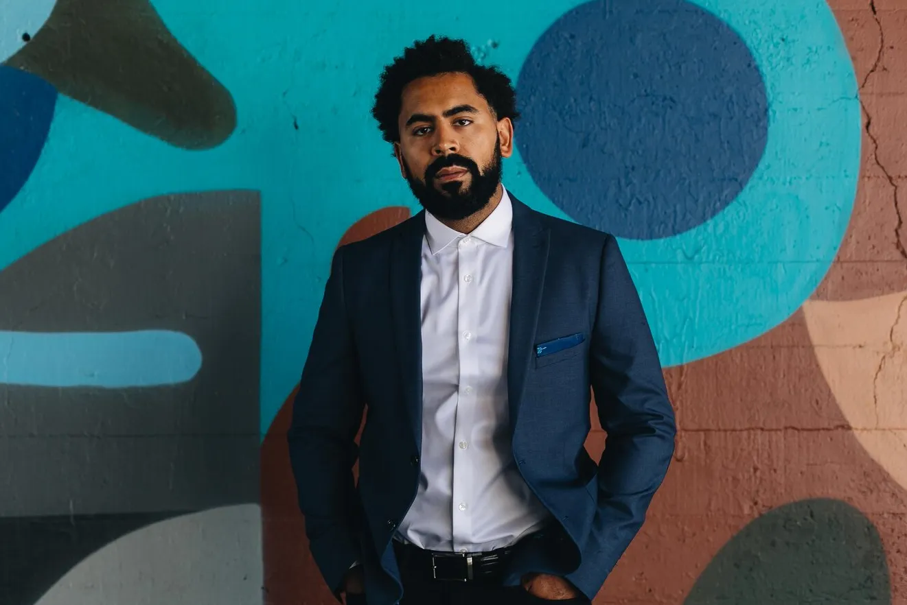 Photo of Rob Eschmann from waist up. He is wearing a blue blazer and white shirt, standing in front of a colorful wall.