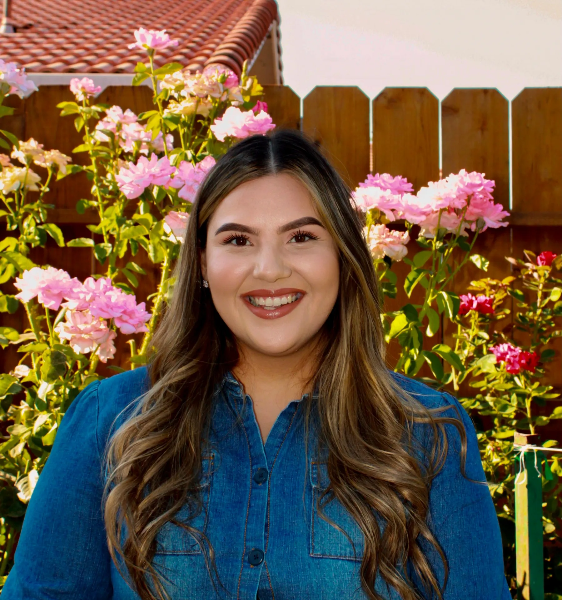 Portrait of Claudia Espinoza-Heredia, a young  female with long brown hair, wearing a jeans jacket against an outdoor  background with pink flowers and the roof of a house