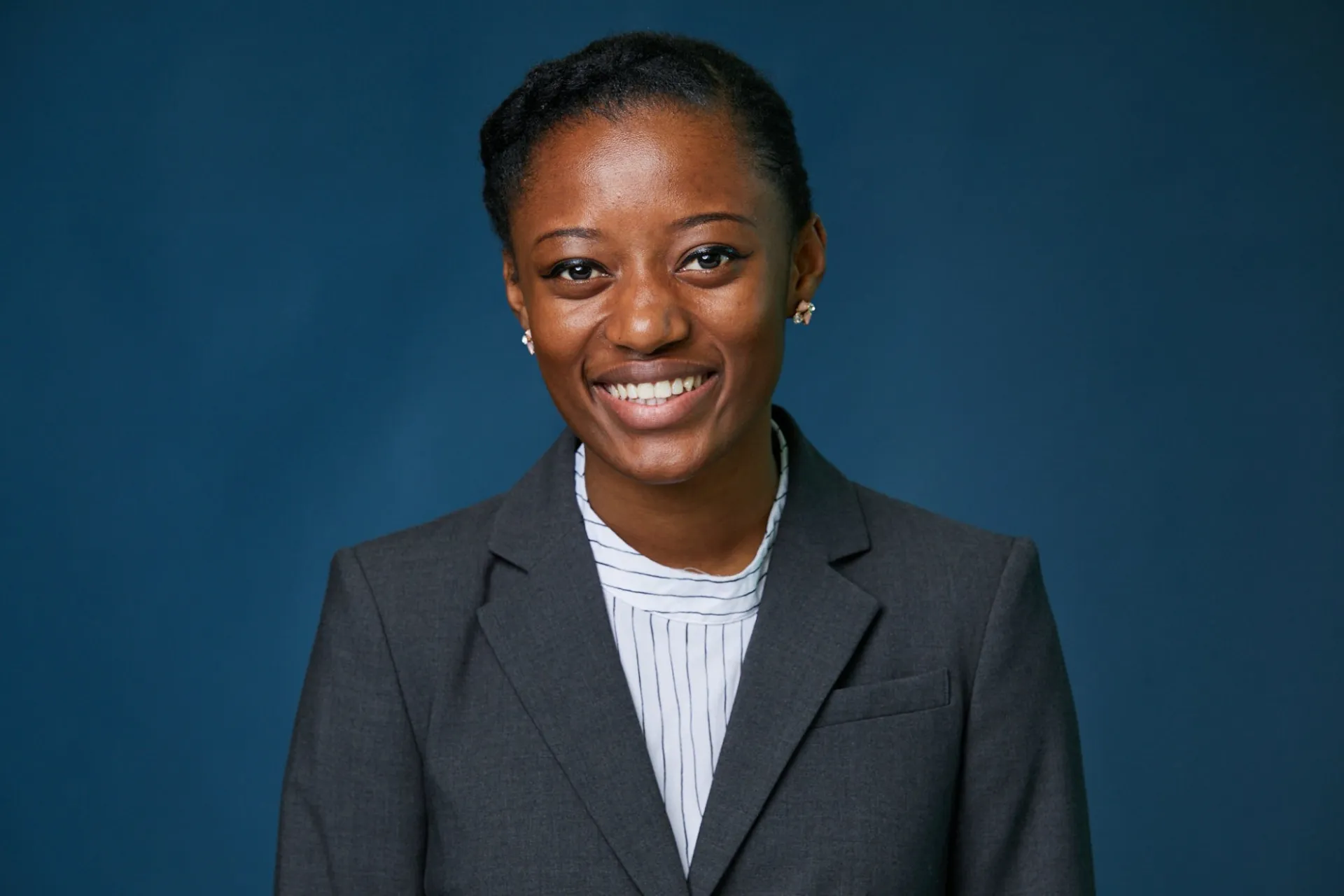 Portrait of Daniela Bushiri, a young female with black hair pulled back, dressed in a grey suit and white stripped shirt, against a dark blue background