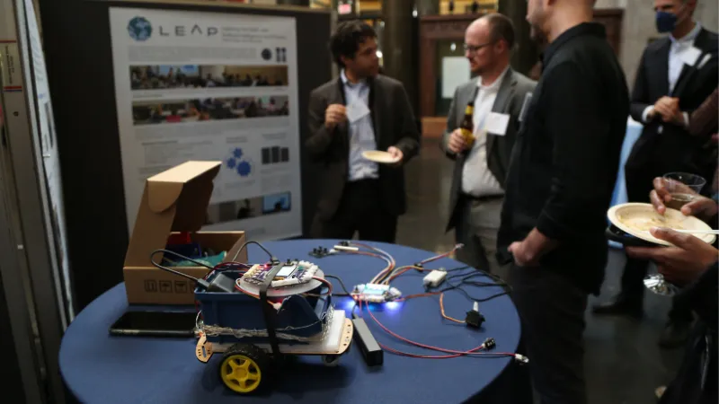 Photo of electronic wheeled vehicle with wires; three men are speaking in the background