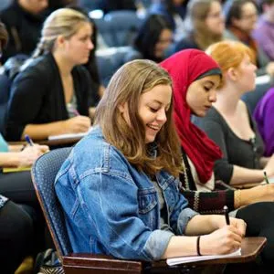 A group of young women at desks in an auditorium, taking notes with paper and pen. 