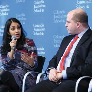 Woman holding a microphone on stage speaks to a man seated beside her.