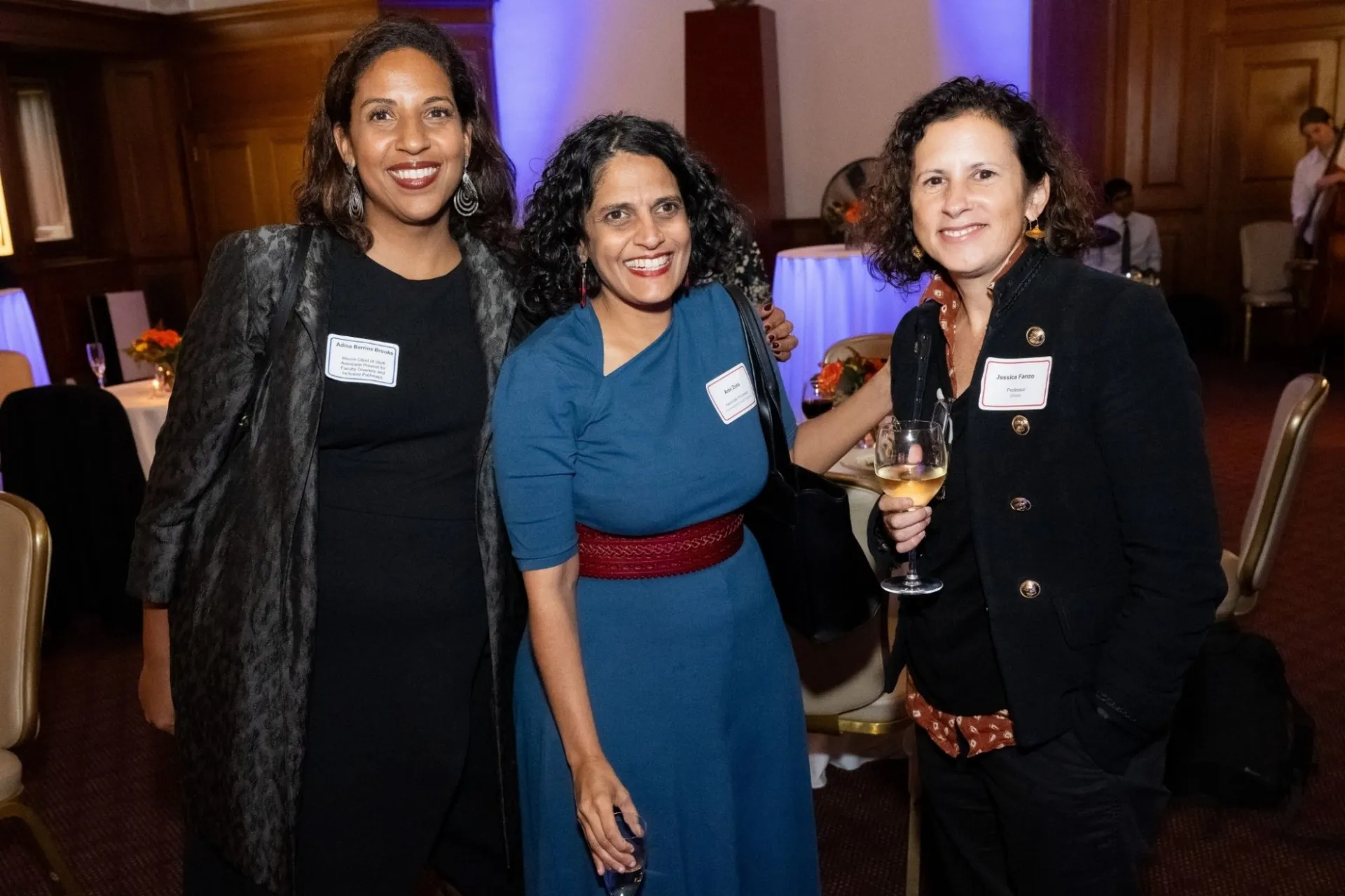 Three women standing together and smiling at a cocktail reception in a room with musicians and tables with seating and orange flowers