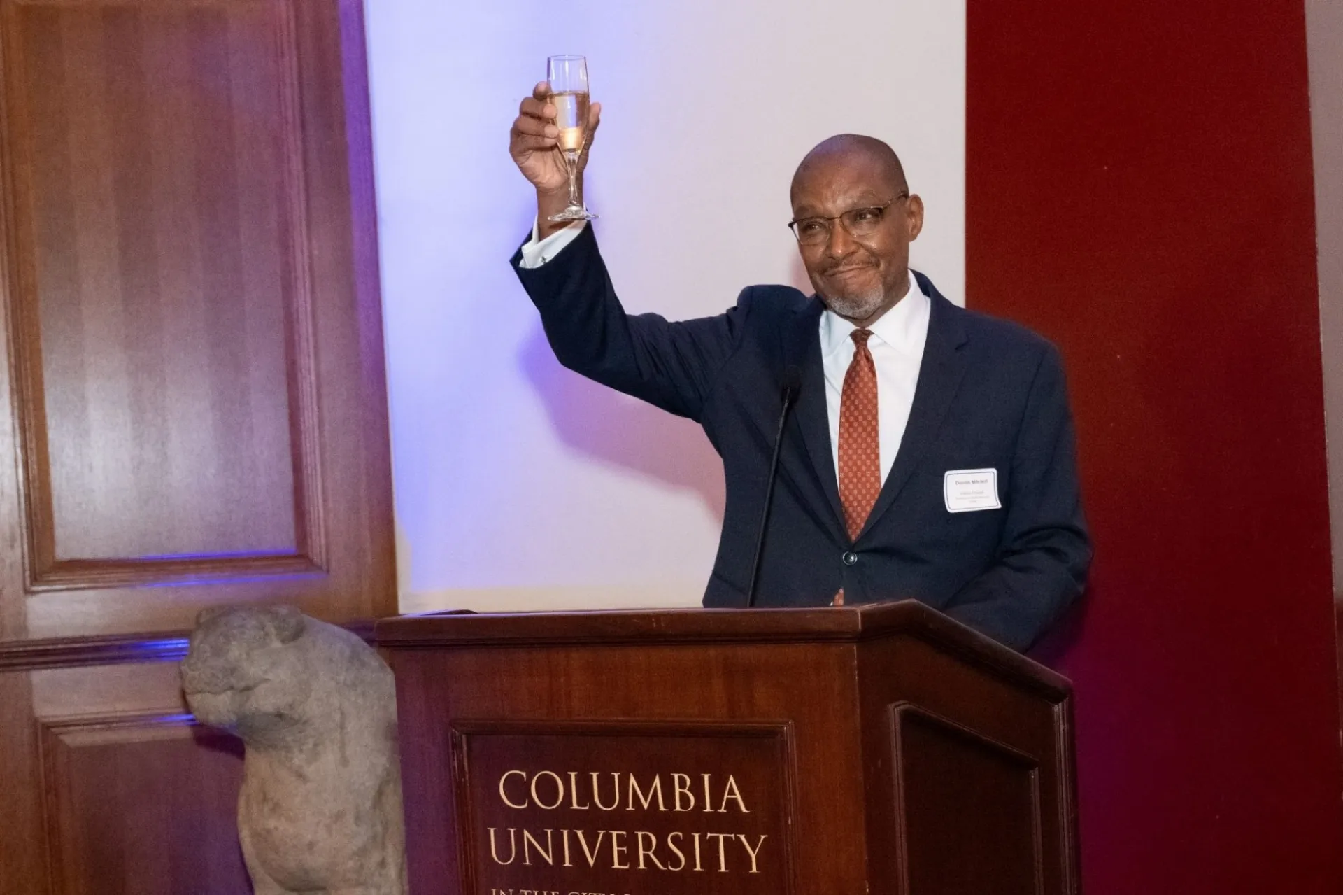 Man in a suit and tie stands at a podium, raising a champagne glass